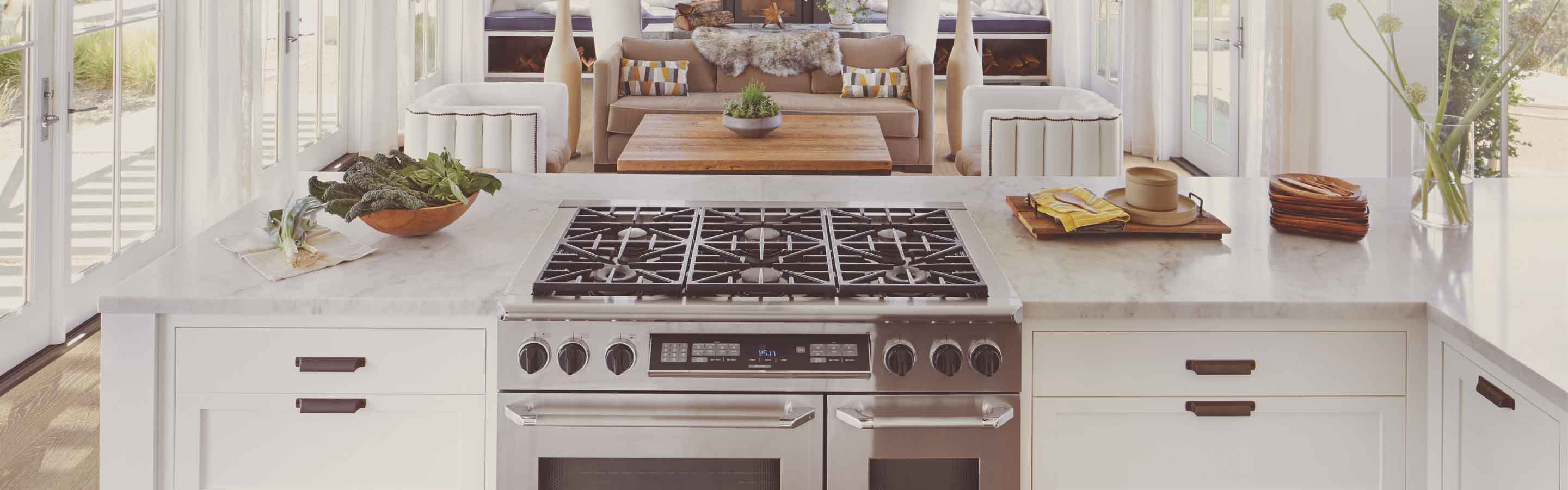 Modern kitchen with marble countertop, stainless steel stove, fresh vegetables, wooden bowl, cutting board, vase with flowers, white cabinets, and an open-concept living area in the background.
